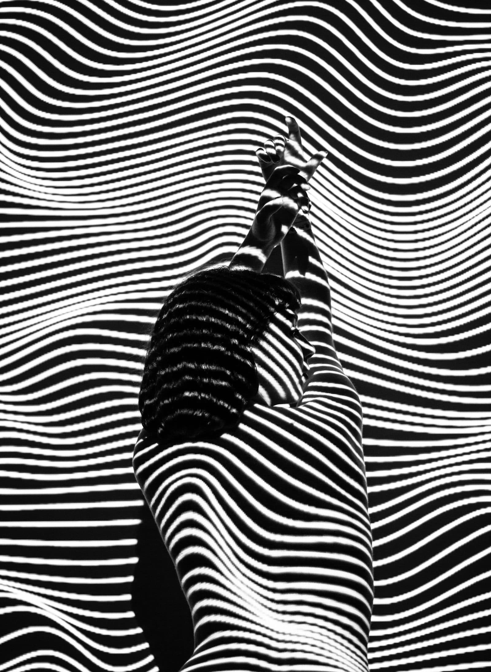 Black and white photograph of a woman seen from behind with her arms raised, covered in rippling stripes of projected light.